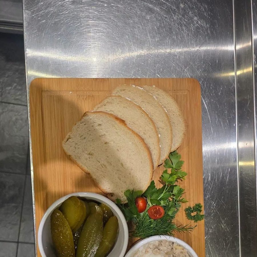 Sliced bread on a wooden board with a bowl of pickled gherkins and a bowl of pale spread.