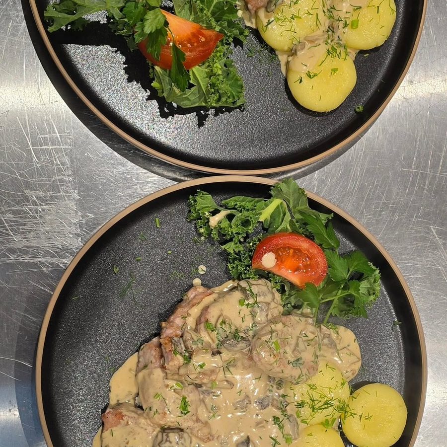Two dark plates of pork in a creamy mushroom sauce with boiled potatoes, tomato and greens.