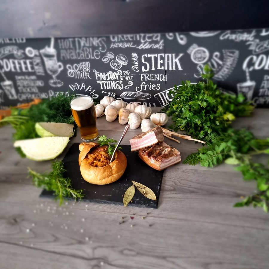 Żurek served in a hollowed bread bowl on a slate board, with a glass of beer and skewers of bacon and gherkin.