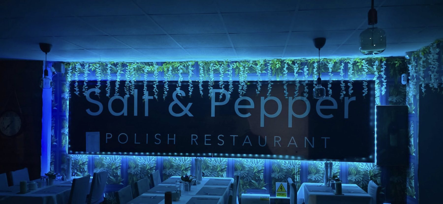 The dining room after dark: white-clothed tables and timber chairs beneath a lit Salt & Pepper Polish Restaurant sign.